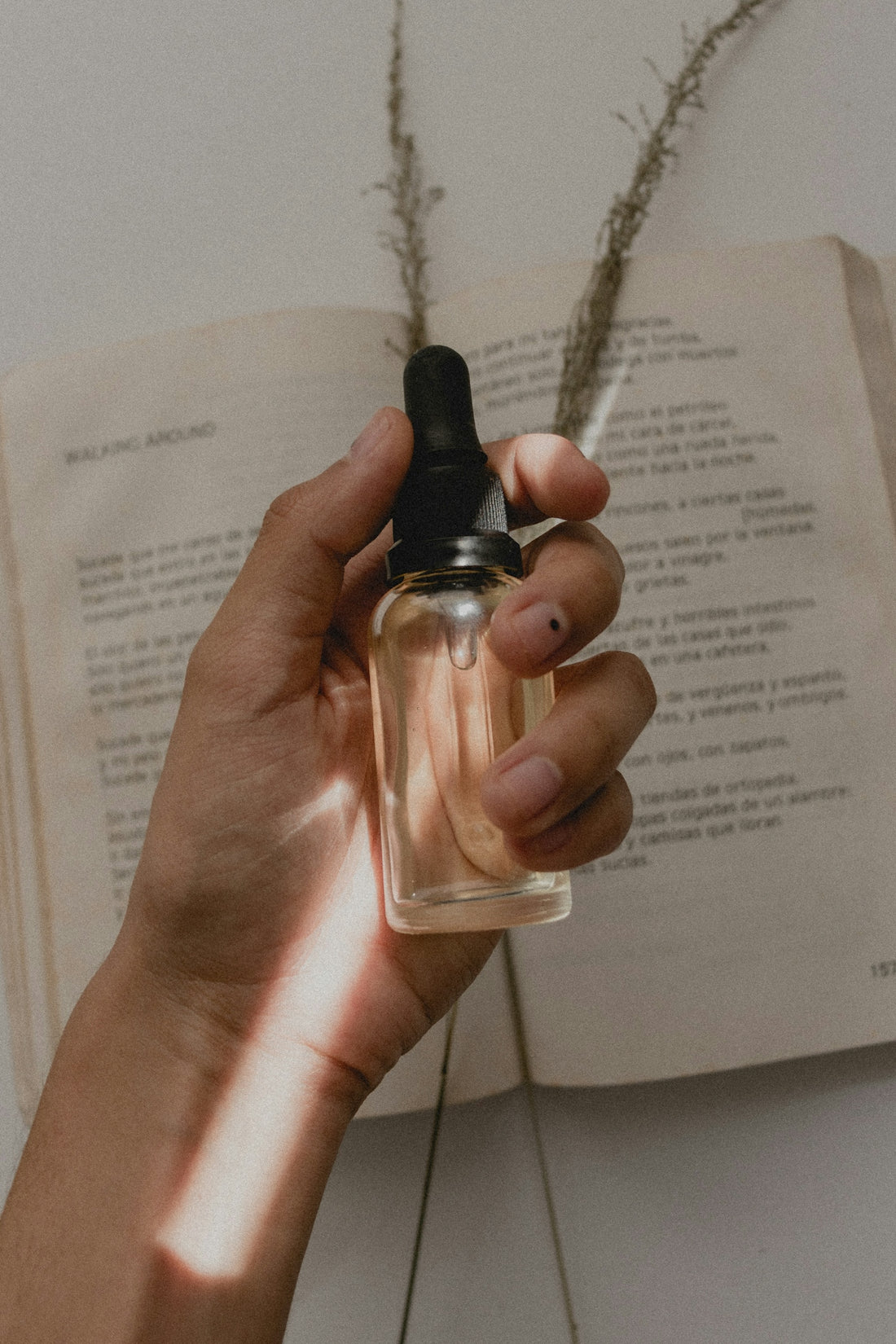 Hand holding a clear glass dropper bottle over an open book for Editorial Navigating The Complex Skincare Market